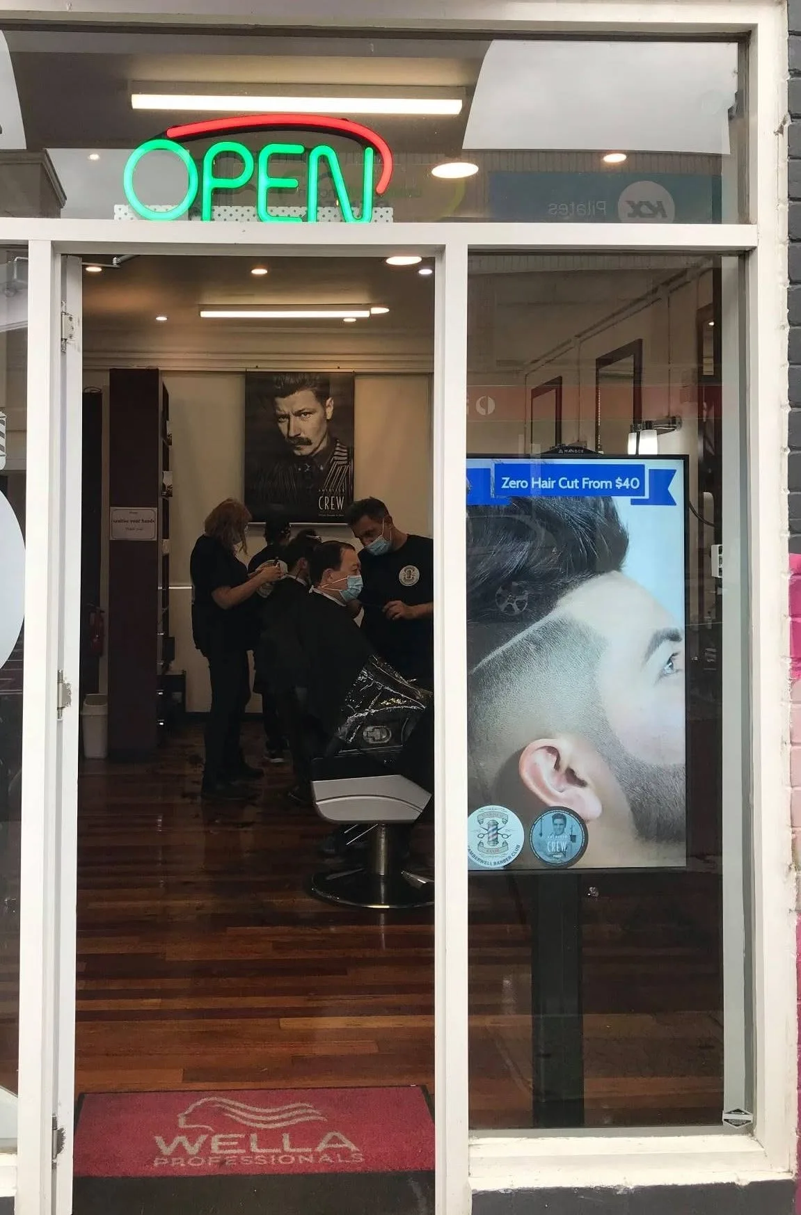 Camberwell Barber Club shopfront — green OPEN neon, barbers at work inside, Wella mat on timber floor